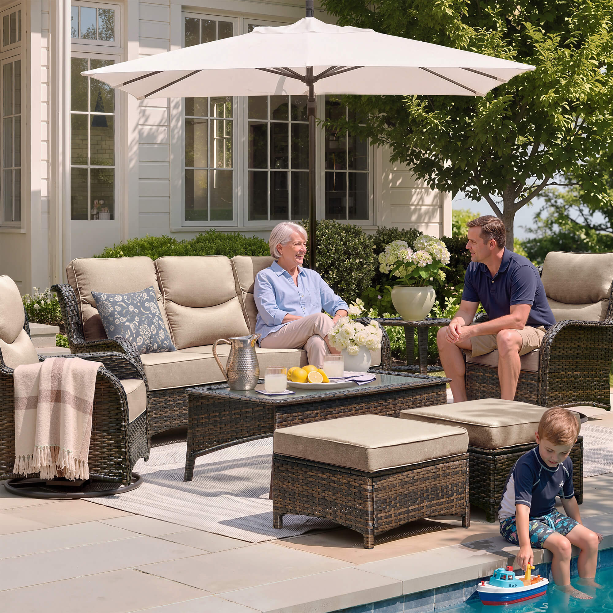 Outdoor patio conversation set with beige cushions and dark wicker furniture, shown in a residential backyard setting with two adults and a child near a pool under a large patio umbrella.